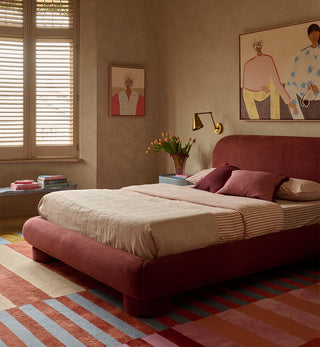 A styled bedroom featuring a prominent dusty rose upholstered bed frame with cream and striped bedding, set against warm beige walls with decorative art, a window with blinds, and a striped rug.