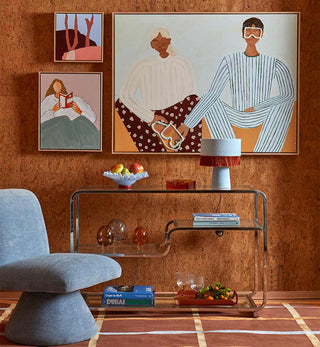 A styled interior scene with a large framed art print on a textured orange-brown wall above a glass console table. The artwork depicts two stylized seated figures, and the room also includes a blue upholstered chair, wall art, a lamp, books, and decor.
