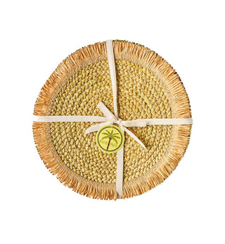 A round woven straw placemat in natural tan and olive tones, tied with a cream ribbon and a small palm tree logo medallion, on a solid white background.