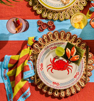 A styled outdoor dining table setting featuring a white ceramic plate with a red crab illustration and the word 'Crabe' printed around the rim, surrounded by a woven placemat. A striped yellow and red napkin with 'Paradis' text, dried orange slices, a glass of citrus juice, and other tableware are arranged on a red and blue striped tablecloth.