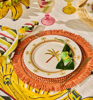 A styled table setting featuring a white ceramic plate with a gold palm tree design, layered over a vibrant orange fringed placemat. The setting includes a yellow and green patterned napkin, gold-toned flatware, and tropical-themed glassware on a white tablecloth.