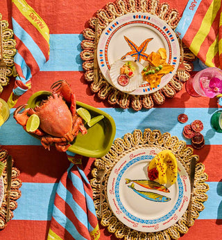 A vibrant beach-themed table setting featuring striped red and blue tablecloths, woven placemats, and matching striped napkins. Two white plates with blue and orange illustrations of starfish and fish are decorated with 'Bienvenue' and garnished with lemon.