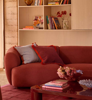 A modern terracotta red upholstered sofa in a warm living room setting, styled with two decorative throw pillows, a glass coffee table, and a wooden bookshelf in the background.