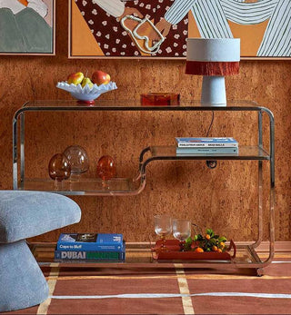 A styled interior scene with a large framed art print on a cork-textured wall above a glass console table. The artwork depicts two seated figures in striped and polka-dot clothing, with two smaller abstract/painting-style prints above.