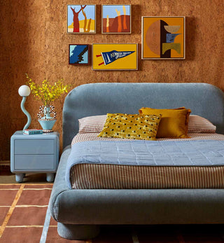 A styled bedroom scene featuring a low-profile bed with a rounded headboard in textured blue-gray fabric, dressed with striped bedding, a mustard yellow pillow, and a patterned yellow accent pillow. The room has a warm brown cork-textured wall decorated with six framed abstract and landscape art prints.
