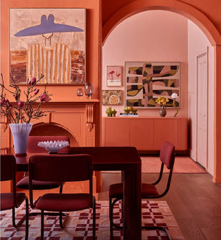 A warm, mid-century modern dining room with terracotta orange walls, a matching sideboard and fireplace, dark wood dining table and chairs, and colorful abstract wall art.