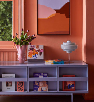 A mid-century modern lavender-gray wooden media console shelf styled in a warm terracotta room. The console features open cubbies and a top surface decorated with a pink tulip vase, a colorful abstract painting, a gray lamp, and books.